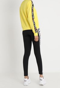 Sudadera amarilla con rayas de logo en blanco y negro en las mangas, combinada con leggings negros que tienen acentos de logo en los laterales y zapatillas blancas.