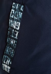 Marinblått tyg med ett texturerat patch med en kontrasterande grafisk design i vitt och ljusblått, broderat längs en kant.