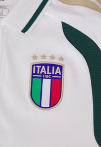 Hvit polo skjorte med grønne detaljer, med et brodert skjoldlogo med blå, grønne, hvite og røde vertikale striper, samt fire gullstjerner.