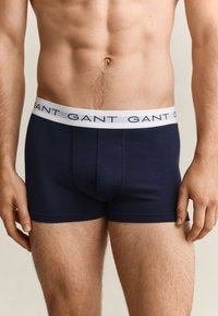 Buste masculin portant un boxer bleu marine avec une ceinture blanche portant le logo de la marque GANT en lettres bleu foncé.