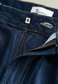 Donkerblauwe denim jeans met gouden stiksels, voorzien van een vierkante metalen knoopsluiting en een ritsgulp. Etiket leest "N. Premium Collection."
