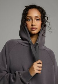 Bluza z kapturem w ciemnoszarym kolorze, o luźnym kroju, z prążkowanymi mankietami i sznurkiem ze metalowymi oczkami. Gładka tekstura.