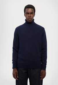 Maglione dolcevita blu navy realizzato in tessuto a maglia, con colletto e polsini a coste, dal taglio rilassato. Abbinato a pantaloni in denim scuro.