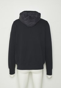 Felpa blu scuro con cappuccio trapuntato e staccabile. Presenta maniche lunghe, polsini a costine e una vestibilità rilassata. Texture liscia e colore uniforme.