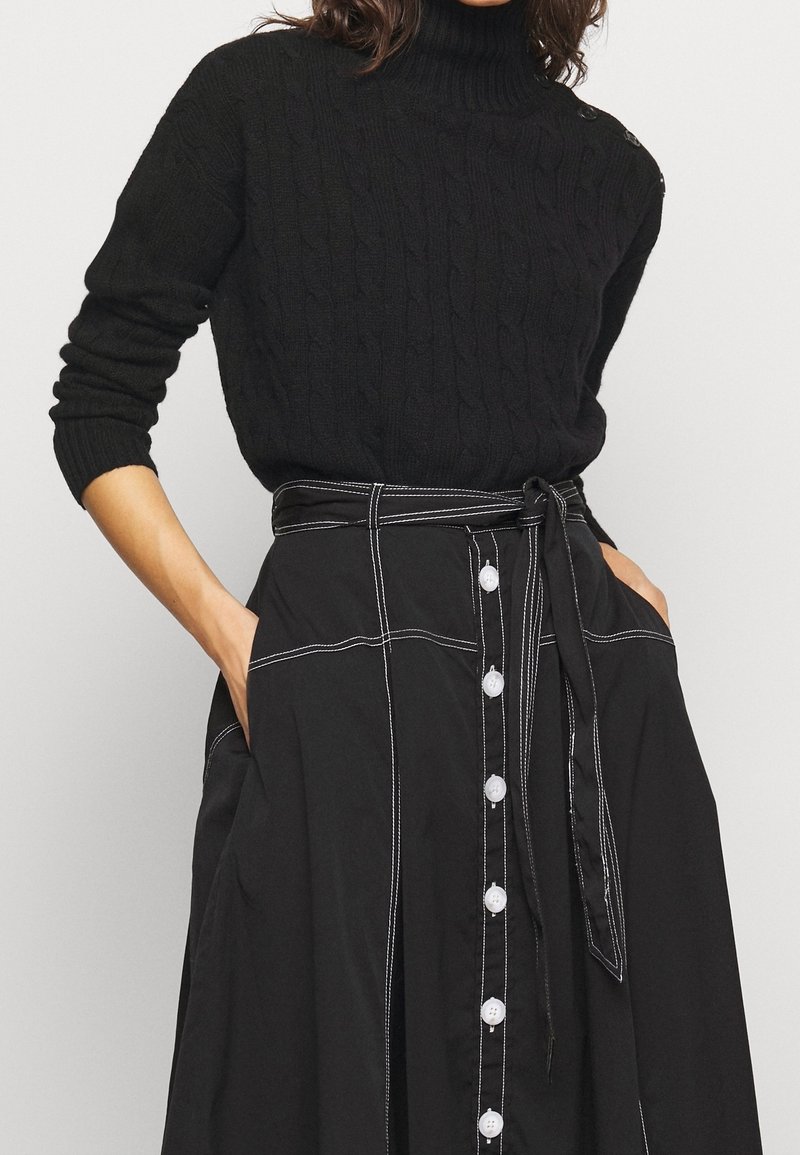 Femme portant un pull à col roulé noir en maille torsadée, rentré dans une jupe noire taille haute à boutons blancs et ceinture nouée, les mains dans les poches.