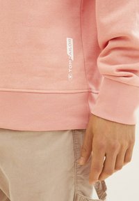 Jasnoróżowa bluza z ribbowanymi mankietami i dołem, z małym białym logo z boku. Miękki, teksturowany materiał bawełniany.