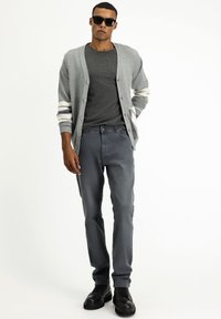 Cárdigan gris con rayas blancas, camiseta gris oscuro, jeans grises y botas negras. El modelo lleva gafas de sol y está de pie contra un fondo liso.