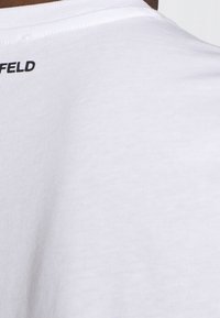Bílý bavlněný tričko s kulatým výstřihem a krátkými rukávy; na zadní části krku je vytištěn černý nápis "FELD". Hladká textura.