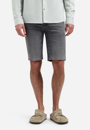 Man draagt een lichtgekleurde overhemd met knopen, grijze spijkershorts en beige instappers, staat met ontspannen handen langs het lichaam.