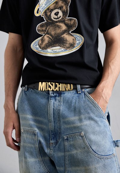 Must värvi T-särk karu graafikuga, millele on sobitatud sinised teksapüksid, millel on kerge pleekimine, ja must vöö, millel on kuldne "MOSCHINO" pandla.