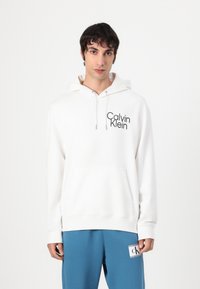 Balta gobtuvė su raišteliais ir priekinėmis kišenėmis, turinti juodą "Calvin Klein" logotipą. Lygus audinys, atsipalaidavęs siluetas. Suderinta su mėlynomis sportinėmis kelnėmis.