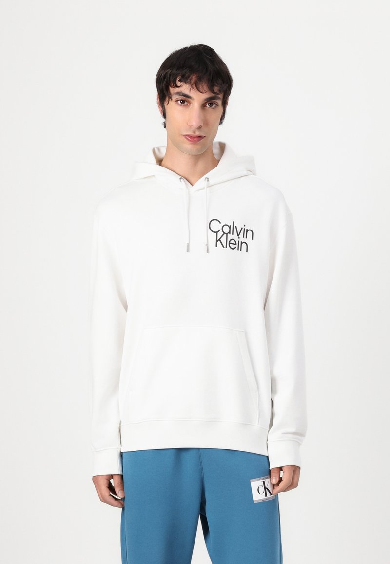 Balta gobtuvė su raišteliais ir priekinėmis kišenėmis, turinti juodą "Calvin Klein" logotipą. Lygus audinys, atsipalaidavęs siluetas. Suderinta su mėlynomis sportinėmis kelnėmis.