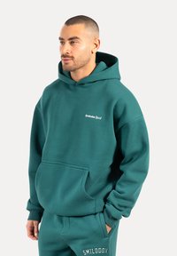 Zielona polarowa bluza z kapturem, z kieszenią kangurką, ściągaczami przy mankietach oraz haftowanym logo. Luźny krój i miękka tekstura zwiększają komfort.