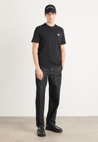 Zwarte katoenen t-shirt met een klein logo, gecombineerd met donkerdenim jeans. Model draagt een zwarte pet en zwarte schoenen, staand op een lichte vloer.