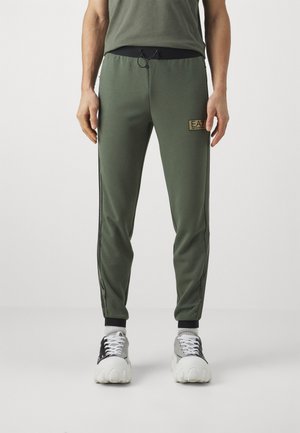 Pantaloni sportivi verdi con un design aderente, vita elastica e dettaglio con cordoncino. Presentano strisce nere sui lati e un patch con logo sulla gamba.