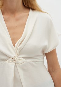 Blusa bianca con scollo a V profondo e maniche corte. Presenta un dettaglio a nodo torcigliato sulla parte anteriore, texture liscia e vestibilità morbida.