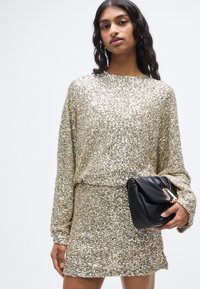 Φόρεμα από χρυσές παγιέτες με χαλαρή εφαρμογή, μακριά μανίκια και στρογγυλή λαιμόκοψη, συνδυασμένο με μαύρο clutch με χρυσή διακοσμητική λεπτομέρεια.