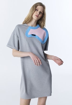 Jonge vrouw die een losse grijze jurk draagt met korte mouwen en een gebreide roze en blauwe patch op de schouder tegen een witte achtergrond.