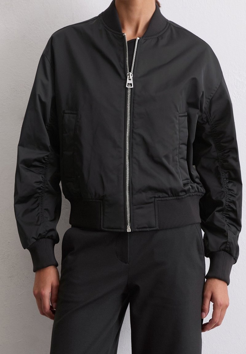 Personne portant une veste bomber noire zippée avec poignets et ceinture côtelés, associée à un pantalon noir, sur un fond clair uni.
