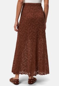 Jupe maxi en dentelle brune texturée, ornée de motifs floraux et d'une taille cintrée. Matériau léger et silhouette fluide.