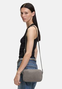 Graue Wildleder-Crossbody-Tasche mit glatter Textur, rechteckiger Form und verstellbarem Riemen, über einer schwarzen Bluse und Jeans an der Schulter getragen.