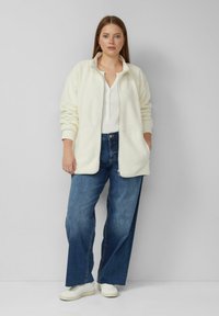 Kurtka z polaru w kolorze ecru z zamkiem i kieszeniami, noszona na białej bluzce, zestawiona z niebieskimi szerokimi jeansami i białymi trampkami.