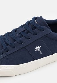 Námorně modré plátěné tenisky s námořnickými tkaničkami, bílé vyšívané logo na straně a silná bílá gumová podrážka.