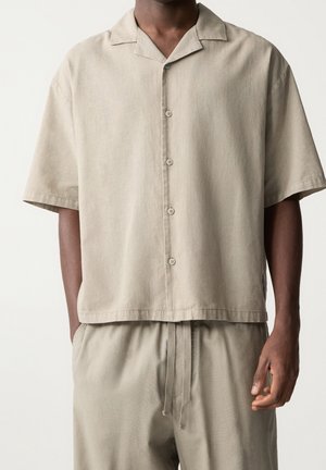 Homme portant une chemise ample beige à manches courtes boutonnée et un pantalon assorti à cordon, mains détendues sur les côtés.