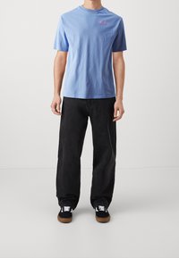 Světle modré tričko s krátkým rukávem a vyšitým růžovým textem na hrudi, doplněné volnými černými džíny a černými teniskami s bílými tkaničkami.