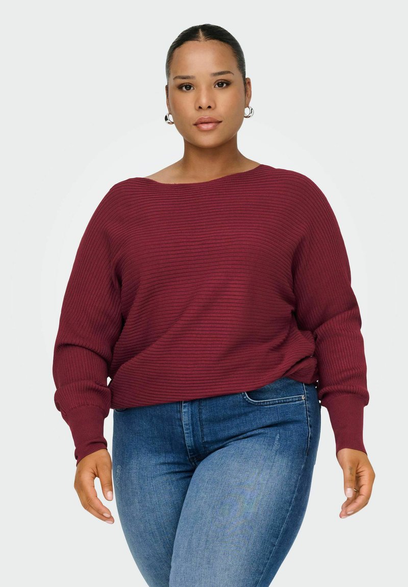 Vrouw met donker haar in een knot, gekleed in zilveren ringoorbellen, een ribbelige donkerrode trui en blauwe jeans tegen een effen lichte achtergrond.
