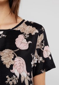 Haut en tissu noir avec broderies florales en rose doux et beige, manches courtes et encolure ronde avec une coupe décontractée.