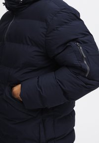 Mørkeblå puffjakke med en quiltet design, lynlåslukning ved manchetterne og sidelommer. Har en høj krave og en glat tekstur.