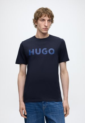 Jonge man met krullend haar die een donkerblauw T-shirt draagt met "HUGO" in blauwe letters en een blauwe spijkerbroek tegen een effen lichte achtergrond.