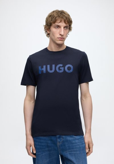 Млад мъж с къдрави коси, носещ тъмносиня тениска с надпис "HUGO" в сини букви и сини дънки на светъл едноцветен фон.