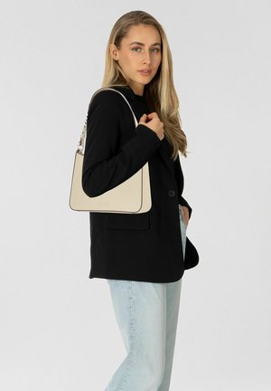 Mujer rubia con blazer negro y jeans azul claro sostiene bolso beige de hombro, posando frente a un fondo liso de color gris claro.