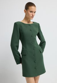 Frau in einem eng anliegenden, dunkelgrünen Langarmkleid mit Knöpfen auf der Vorderseite und aufgesetzten Taschen, die vor einem schlichten, hellen Hintergrund steht.