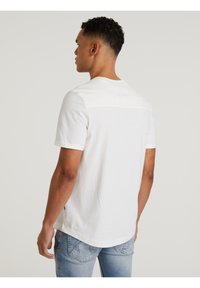 Weißes Baumwoll-T-Shirt mit kurzen Ärmeln, Rundhalsausschnitt und einem Kontraststoff-Panel am oberen Rücken. Getragen mit hellblauen Jeans.