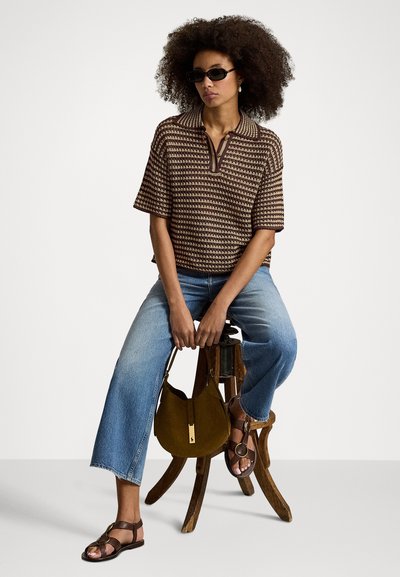 Ženska z kodrastimi lasmi, ki nosi sončna očala, rjavo kratko-sleeve majico z vzorcem, modre kavbojke in sandale, sedi na lesenem stolčku in drži rjavo torbico.