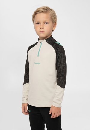 Blond dreng iført en langærmet sportstop med sorte mønstrede ærmer og beige front, stående mod en ensfarvet baggrund.