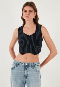 Izgriezts melnā pinstrīpu veste ar V-veida kakla izgriezumu, ar priekšējiem pogām un platām plecu siksnām, kopā ar gaiši ziliem augstiem džinsiem.