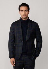 Uomo che indossa un completo a quadri verde scuro e blu navy con pantaloni abbinati e un maglione nero a collo alto, in piedi con una mano in tasca.