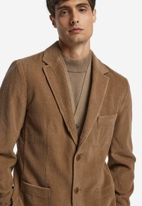 Brauner Cord-Blazer mit Spitzkragen, Brusttasche und Zwei-Knopf-Verschluss. Über einem passenden Pullover getragen für einen gestuften Look.
