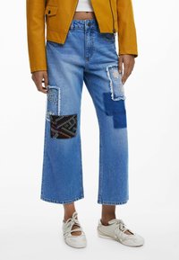 Hellblaue Denim-Wide-Leg-Jeans mit ausgefranzten Patches, die auf beiden Oberschenkeln mit bestickten und gemusterten Stoffakzenten versehen sind.