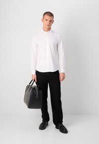 Hvit skjorte med knapp, svarte bukser, svart lærveske og sko med tykke såler. Modellen står mot en ensfarget bakgrunn.