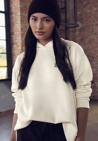 Felpa crema con tasca a marsupio e polsini a costine, con un piccolo logo nero. Abbinata a pantaloni a righe sottili scuri e un berretto di maglia marrone.