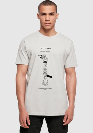Ung mand iført en hvid t-shirt med et diagram over en kaffemaskine og teksten, der definerer "depresso" som at løbe tør for kaffe.