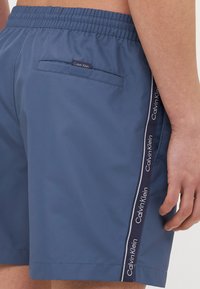Mėlyni maudymosi šortai su elastingu juosmeniu, turintys galinę kišenę ir šoninį juostelės apdailą su "Calvin Klein" ženklu.