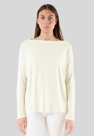 Donna con capelli lunghi castani indossa una camicia larga a maniche lunghe color avorio e pantaloni bianchi, in piedi davanti a uno sfondo grigio uniforme.