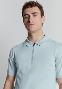 Lichtblauwe polo met korte mouwen en een ritskraag, gemaakt van zachte stof. Bevat ribbelboorden bij de mouwen en subtiele stiksels.