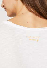Weißes T-Shirt mit entspanntem Ausschnitt und strukturiertem Stoff, mit "Passion Love Heart" und "MOS MOSH" in Gelb auf der Rückseite bedruckt.